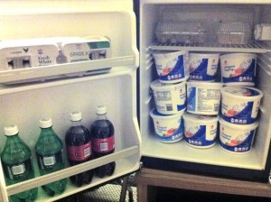 A hotel room fridge, with some sodas in the door and nine tubs or whipped topping defrosting in the main section